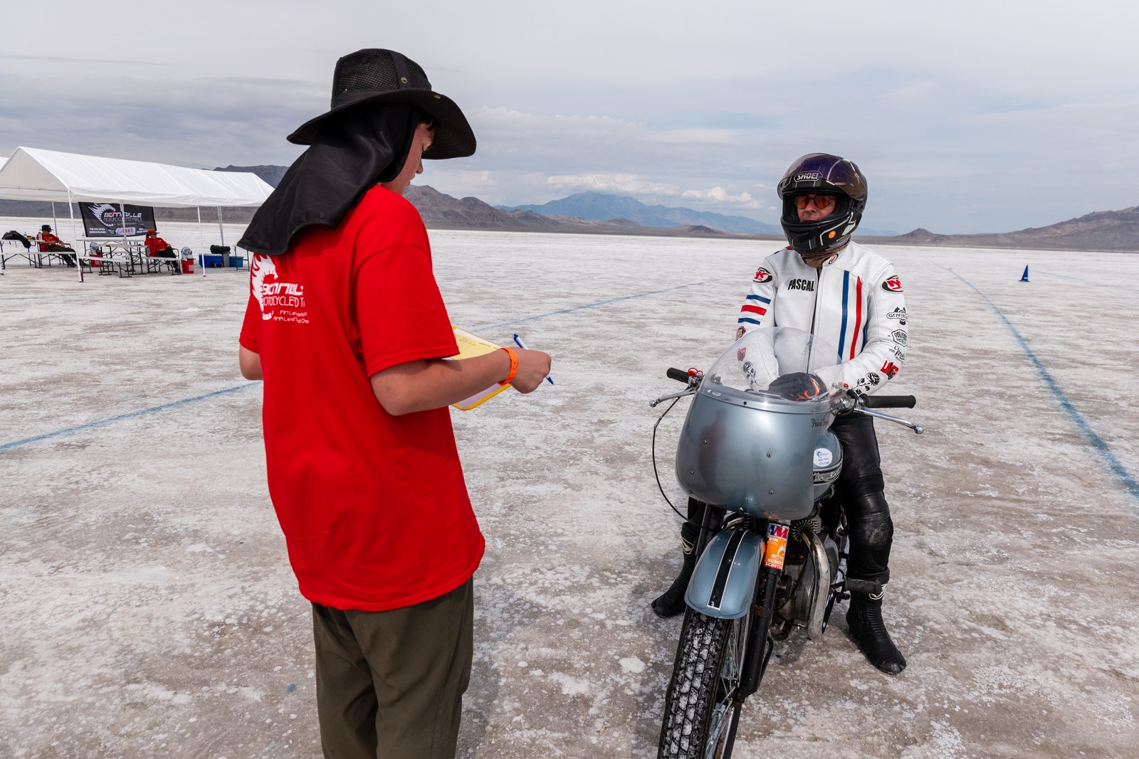 A race official in a cowboy hat talking with a rider and a vintage motorcycle on the salt