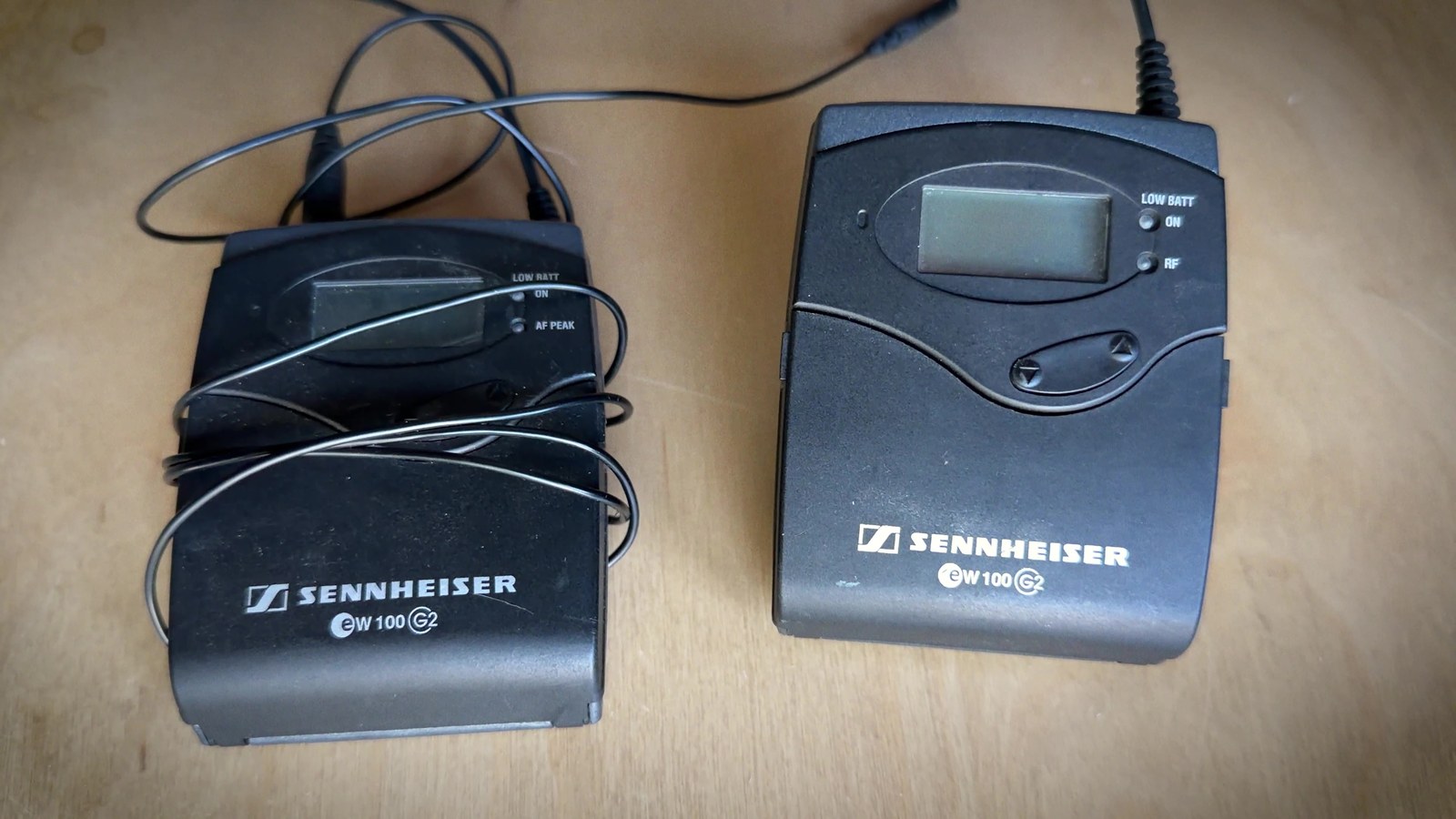 A pair of older Sennheiser EW100 G2 wireless bodypack transmitters on a wooden desk