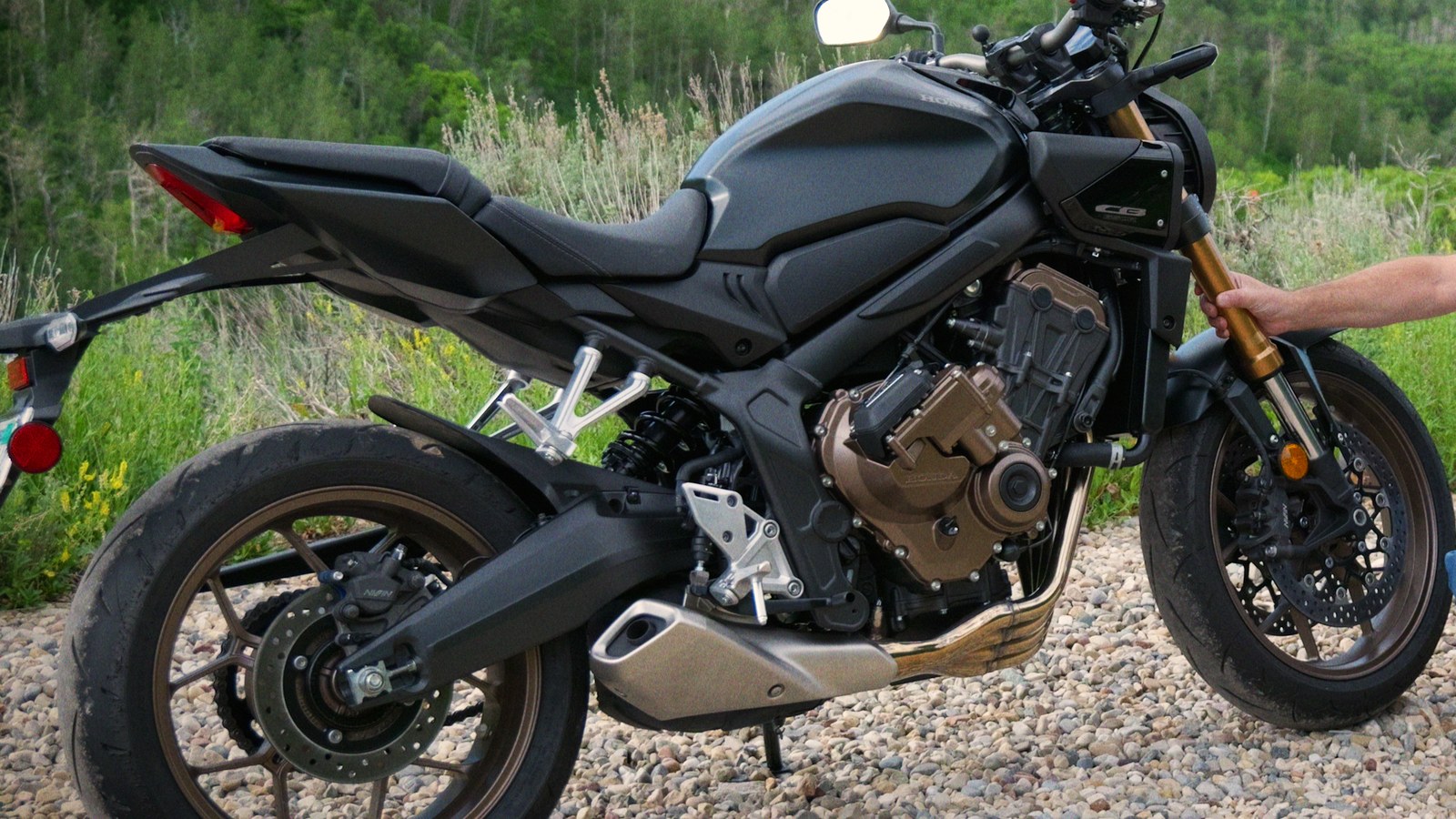 Honda CB650R in Matte Gunpowder Black Metallic parked on a gravel pullout with a green hillside behind